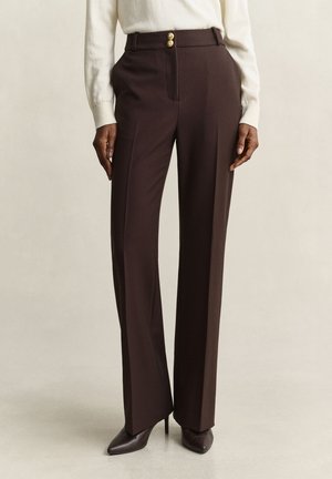 Personne portant un pantalon sur mesure marron foncé taille haute avec des boutons dorés, associé à des talons pointus marron foncé et un pull blanc.