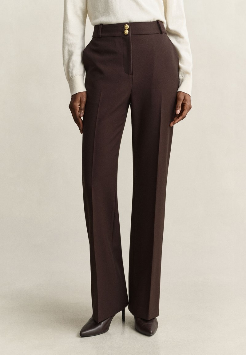 Personne portant un pantalon sur mesure marron foncé taille haute avec des boutons dorés, associé à des talons pointus marron foncé et un pull blanc.