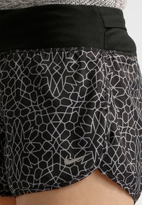 Nike-shortsit musta-harmaalla geometrisella kuviolla, joissa on musta joustava vyötärö ja sivuhalkio liikkuvuuden takaamiseksi. Hopeinen logo korostaa.