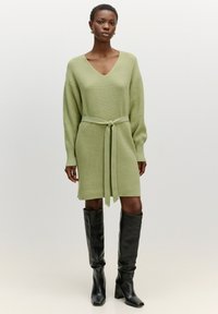 Brede jurk in lichtgroen met een V-hals, lange mouwen, en een strikceintuur in de taille. Gepaard met kniehoge zwarte leren laarzen.