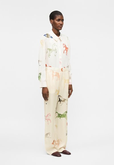 Γυναίκα που φοράει λευκό πουκάμισο με κουμπιά και αντίστοιχο παντελόνι με πολύχρωμα σχέδια αλόγων, συνδυασμένα με σκούρα καφέ παπούτσια, που στέκεται πλαγίως.