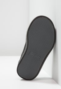 Černá gumová podrážka boty s texturovaným, vroubkovaným povrchem. Obsahuje logo Lacoste, které je viditelné ve středu. Bílý pozadí.