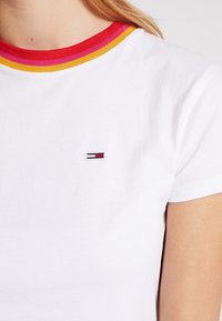 Camiseta blanca de manga corta con cuello acanalado a rayas en rojo, rosa y amarillo, pequeño logotipo rectangular rojo, blanco y azul en el pecho.