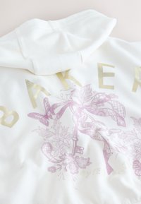 Sweat à capuche blanc avec texte doré "BAKER" et un motif floral rose comprenant une clé et un ruban dans le dos.