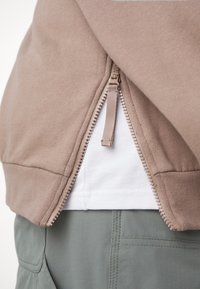 Felpa beige con zip parzialmente aperta sopra una maglietta bianca infilata in pantaloni grigi con cuciture e dettagli delle tasche visibili.