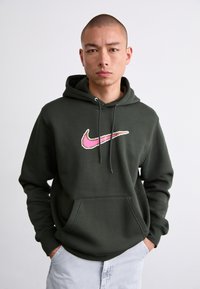 Mørkegrøn Nike hoodie med en frontlomme, justerbar snor og en lyserød swoosh-logo. Glat stoftekstur, afslappet pasform.