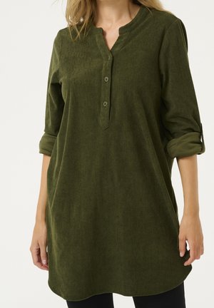 Vrouw draagt een olijfgroene corduroy tuniek met lange mouwen, een drieknopsplacket en opgerolde mouwen.