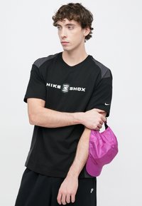 Μαύρο μπλουζάκι Nike Shox με γκρι λεπτομέρειες, με λογότυπο στο μπροστινό μέρος. Ο φορέας κρατάει ένα φωτεινό ροζ καπέλο. Είναι ορατά μαύρα σορτς.