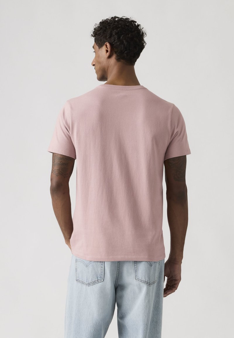 Homme aux cheveux bouclés portant un T-shirt uni rose clair à manches courtes et un jean bleu clair, face à une surface unie, de dos.