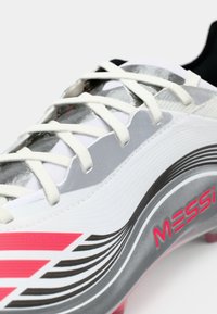 Hvid fodboldsko med grå accenter, der har pink og sorte striber samt "MESSI" branding. Den har en tekstureret overflade og snørebåndsdesign.