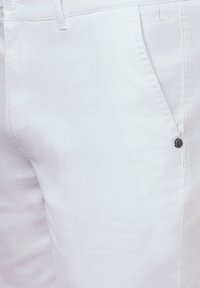 Białe bawełniane spodnie z teksturowanym wykończeniem, z przednią kieszenią i czarnym guzikiem jako akcentem. Subtelne logo wyeksponowane na szwie blisko kieszeni.