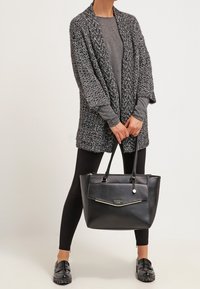 Cardigan a maglia grigia e nera, top a maniche lunghe nero, leggings neri e una grande borsa in pelle nera con accenti dorati.
