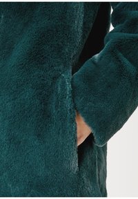 Zielona, sztuczna futrzana kurtka w zbliżeniu, z miękką fakturą, długimi rękawami i boczną kieszenią, podkreślająca puszysty materiał i głębię koloru.