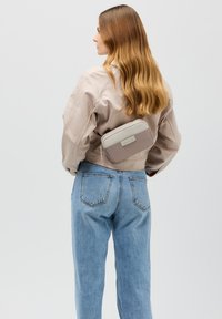 Giacca beige corta sopra jeans blu a vita alta, con una borsa a tracolla zip taupe e grigio chiaro posizionata sulla schiena.
