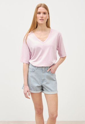 Frau mit langen, glatten Haaren, die ein hellrosa, übergroßes T-Shirt mit V-Ausschnitt und hellblaue, hochgekrempelte gestreifte Jeansshorts trägt, steht mit der Hand in der Tasche.