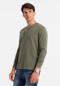 Mann in olivgrünem Langarm-Henley-Shirt und blauer Jeans, der nach links schaut, vor einem schlichten weißen Hintergrund.