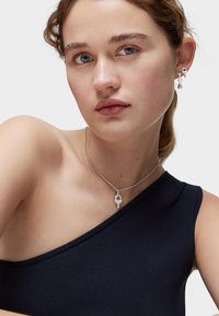 Collar con colgante de candado plateado sobre una cadena delicada, acompañado de pendientes a juego. La modelo lleva una blusa de un solo hombro en color marino. Textura de piel suave visible.