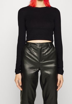 Person mit roten Haaren, die ein schwarzes, geripptes, langärmeliges Crop-Top und hoch taillierte schwarze Kunstlederhosen trägt, steht vor einem schlichten Hintergrund.