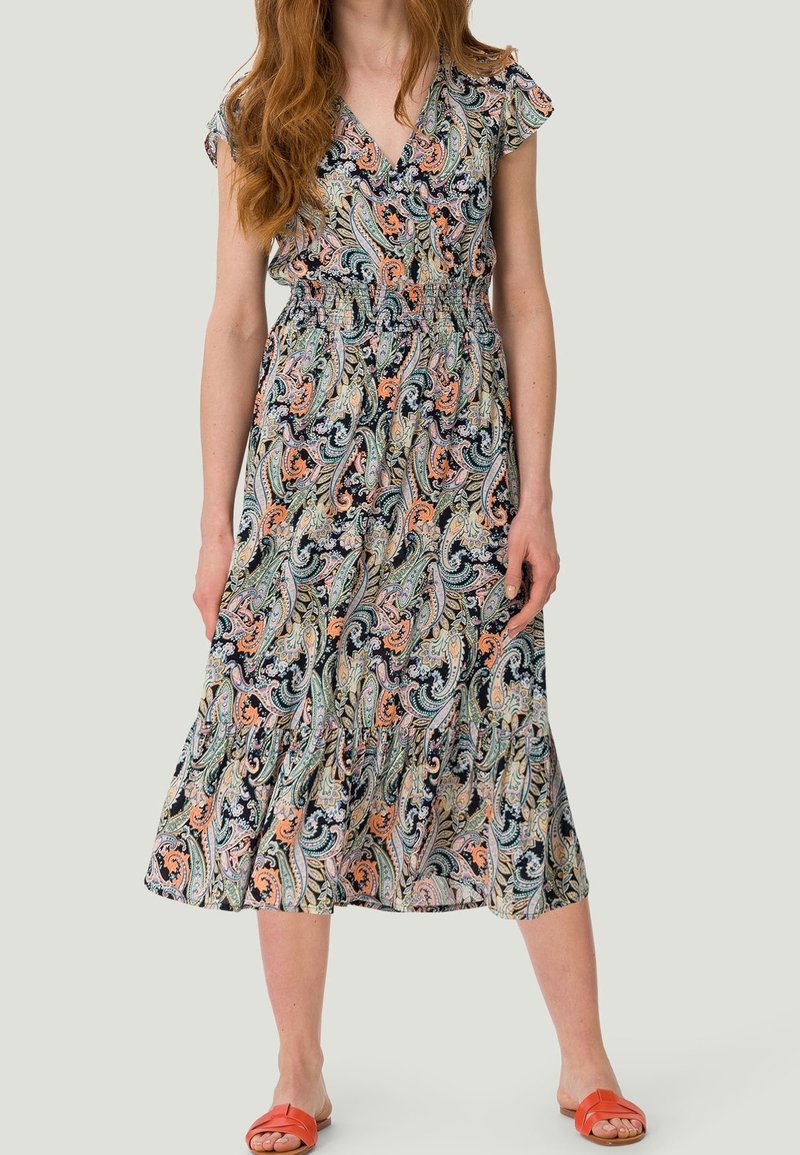 Kvinde iført en farverig paisley-mønstret midi kjole med korte ærmer og orange sandaler, stående mod en ensfarvet baggrund.