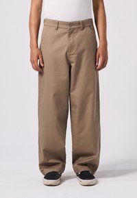Bruine wijde broek van stevig materiaal, met een knoopsluiting aan de voorkant en schuin geplaatste zijzakken. G gedragen met zwarte schoenen.