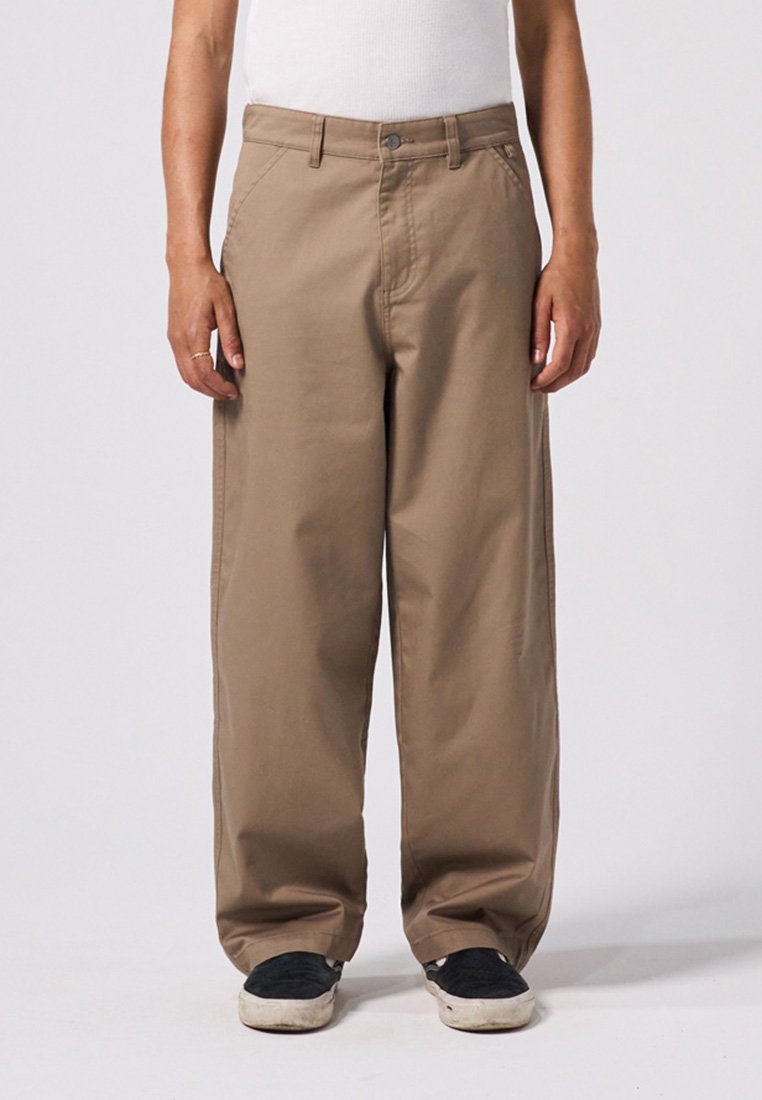 Bruine wijde broek van stevig materiaal, met een knoopsluiting aan de voorkant en schuin geplaatste zijzakken. G gedragen met zwarte schoenen.