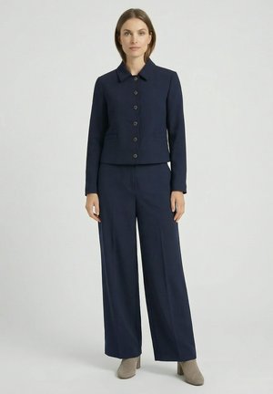 Femme debout portant une veste boutonnée bleu marine avec un pantalon large assorti et des bottines à talons beige sur fond uni.