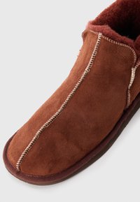 Brown suede slipper featuring a rounded toe, contrast stitching, and a soft fur lining. The sole is a darker brown with a smooth finish.