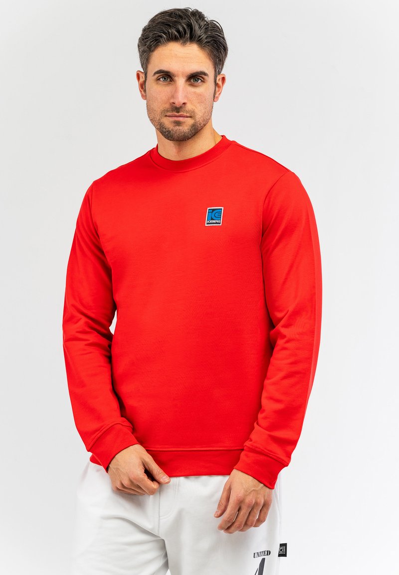 Felpa rossa a maniche lunghe realizzata in tessuto morbido, con una patch logo blu sul petto. Polsini a coste e design a girocollo.