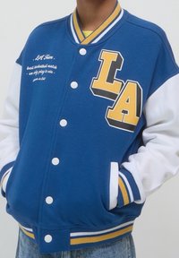 Niebieska kurtka varsity z żółtymi literami „LA”, białymi rękawami, prążkowanym kołnierzem z niebiesko-żółtymi paskami oraz zatrzaskami.
