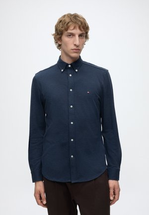 Homme aux cheveux blonds bouclés portant une chemise bleu foncé à boutons et un pantalon sombre sur un fond clair uni.