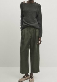 Tummanharmaa neulottu neuletakki, jossa on avara kaula-aukko, yhdistettynä löysiin, oliivinvihreisiin pellavahousuihin. Beiget liukujalkineet täydentävät asua.