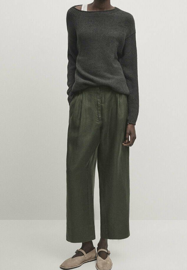 Tummanharmaa neulottu neuletakki, jossa on avara kaula-aukko, yhdistettynä löysiin, oliivinvihreisiin pellavahousuihin. Beiget liukujalkineet täydentävät asua.