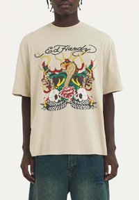 Camisa de algodón beige con un gráfico colorido de serpientes, calaveras y una flor. Corte holgado, con mangas cortas y diseño de cuello redondo.