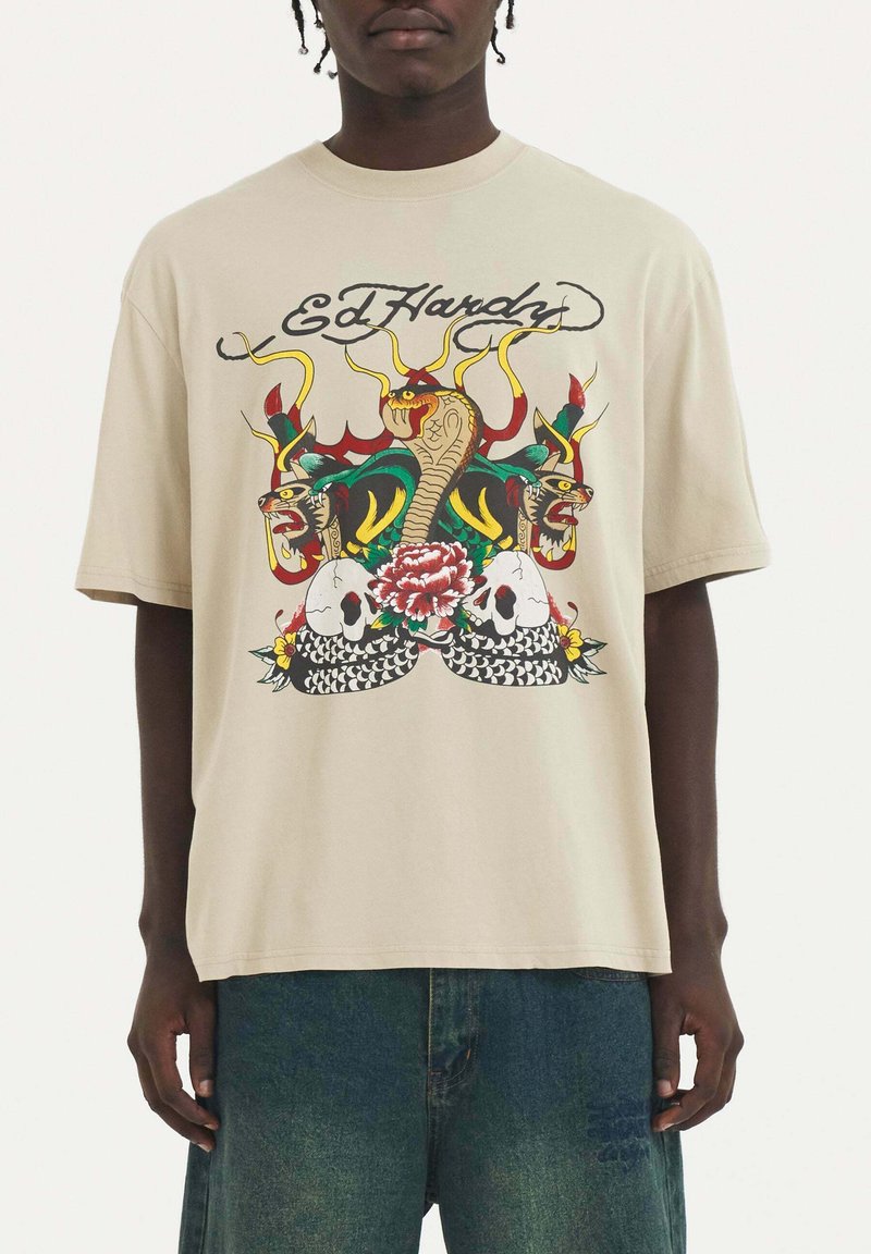 Camisa de algodón beige con un gráfico colorido de serpientes, calaveras y una flor. Corte holgado, con mangas cortas y diseño de cuello redondo.
