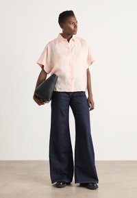Blusa rosa chiaro a maniche corte con bottoni abbinata a jeans a gamba larga blu scuro. Il modello tiene una borsa semi-strutturata nera. Texture liscia, design minimal.