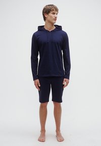 Camisola com capuz leve azul marinho com cordões, combinada com calções compridos até ao joelho a condizer. Textura suave e design ajustado. Modelo descalço.