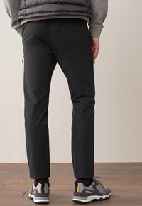 Musta housut, joissa on k slim fit -malli ja vetoketjulliset taskut, yhdistettynä harmaisiin ja keltaisiin vaelluskengiin, puisten lattioiden taustalla.