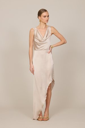 Frau in einem ärmellosen champagnerfarbenen Satinkleid mit Wasserfallausschnitt und asymmetrischem Saum, trägt goldene Ohrringe, Armband und Riemchenabsätze.