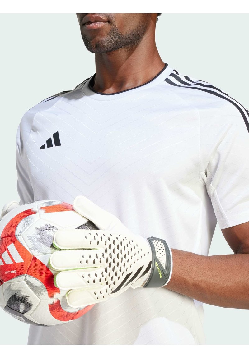 Guanti da portiere in materiale latex bianco, con grip testurizzato e dettagli verdi. Il giocatore tiene un pallone da calcio colorato, indossando una maglia bianca con righe nere.