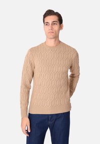 Maglione a maglia beige con un motivo a nido d'ape testurizzato e polsini e orlo a coste. Scollo tondo e maniche aderenti. Indossato con jeans scuri.
