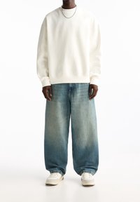 Personne portant un sweat-shirt blanc, un jean bleu clair ample, des baskets blanches et une fine chaîne autour du cou, se tenant devant un fond blanc.