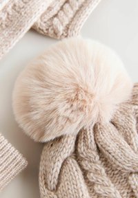Beige gestrickte Mütze mit einem großen, weichen Kunstfell-Pompon oben, ausgestattet mit einem strukturierten Zopfmuster und einem flachen gestrickten Schirm.