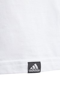 Camiseta blanca de algodón con una textura en la tela, dobladillo recto y una etiqueta negra de Adidas en la esquina inferior izquierda.
