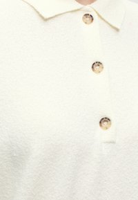 Κοντινό πλάνο σε ύφασμα κρέμας με γιακά και τρία κουμπιά από φολιδωτό φλοιό χελώνας τοποθετημένα σε κάθετη σειρά.