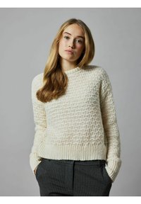 Cremfarbener, strukturierter Pullover mit Rautenmuster, geripptem Saum und Bündchen. Kombiniert mit dunkel gestreiften Hosen.