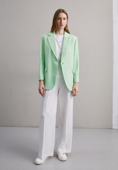 Blazer vert menthe avec un design structuré, fermeture à un bouton et revers, associé à un pantalon large blanc et des baskets.