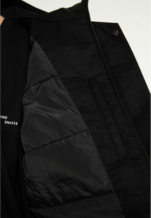 Svart jacka med foder i kviltat material, slät yta och dolda fickor. Designen inkluderar en dragkedja och tryckknappsstängning.