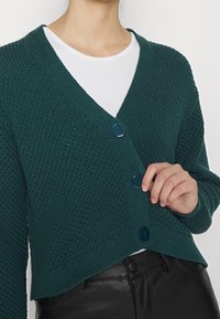 Žena zapínající tmavě zelený texturovaný pletený cardigan přes bílou košili s černými koženými kalhotami, záběr na trup a ruku.