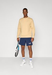 Beige sweatshirt, navy shorts with EA7 logo, white sneakers, blue-accented tennis racket. Simple design, athletic wear, relaxed fit.