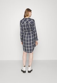 Abito camicia a quadri navy con maniche lunghe, vita stretta, chiusura con bottoni sul retro e orlo asimmetrico, abbinato a stivali bianchi alla caviglia.